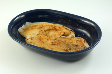 Fresh raw tilapia fish fillets prepared for cooking with spicy rub of chili powder, garlic and cumin in blue oval dish on white wooden table