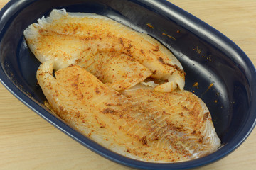 Close up of fresh raw tilapia fish fillets prepared for cooking with spicy rub of chili powder, garlic and cumin in blue dish