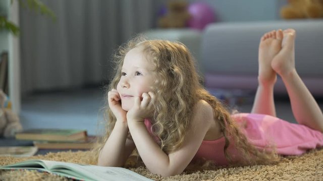 Cute curly-haired girl lying on soft carpet at home and smiling, happy child