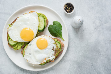 Two toasts with avocado, cucumber and egg on white plate over grey concrete background. Top view with copy space for text