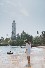 Fototapeta na wymiar Young woman in white dress and hat walking along the beach against the lighthouse