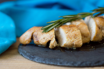 Slices of baked chicken breast served with green rosemary leaves on trendy black slate board with blue napkin, selective focus