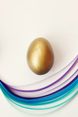 Golden egg on white background, decorated with blue and violet paper stripes