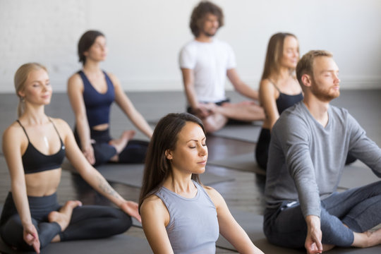 Group of young sporty people practicing yoga lesson with instructor, sitting in Padmasana exercise, meditating with closed eyes, Lotus pose, working out, indoor, students training in club, studio