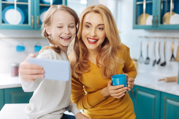 Good mood. Attractive joyful fair-haired slim mother smiling and holding a cup of tea while her daughter taking selfies