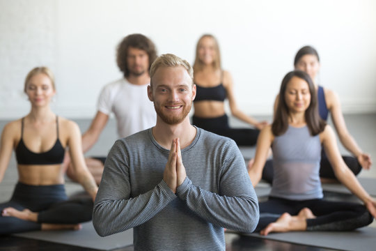 Portrait of young friendly smiling male yoga instructor looking at camera, group of sporty people practicing lotus pose, sitting in Padmasana exercise, working out, students training in club, studio