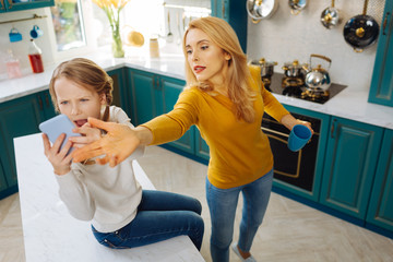 Anxious. Uncontrolled fair-haired slim girl holding a phone and crying while her mom taking it away