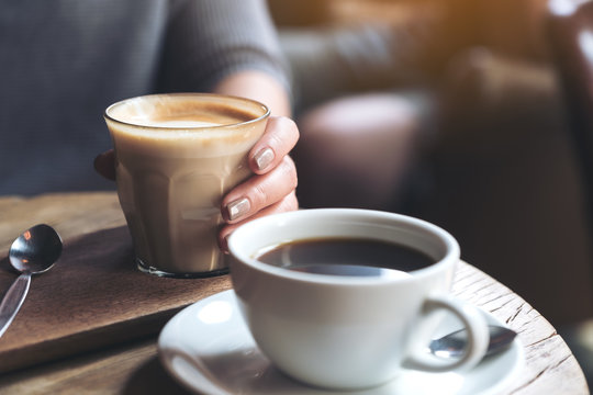 Closeup image of Americano coffee cup and woman's hand holding latte coffee cup on vintage wooden table in cafe