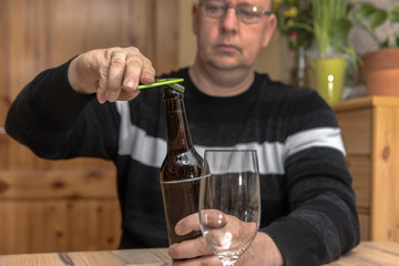 A man (45-55) sits at the table and opens a bottle of beer. Concept: alcohol abuse or drinking