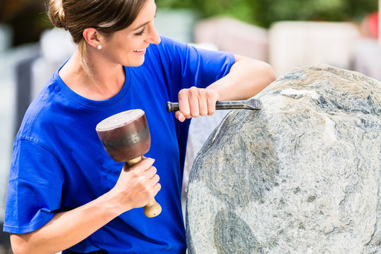 Female stonemason working on boulder with sledgehammer and iron