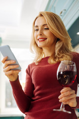 Resting. Pretty joyful blond young woman smiling and typing on her phone while drinking some red wine and relaxing