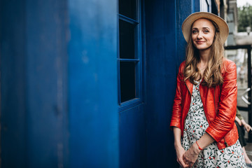 Obraz na płótnie Canvas Attractive portrait of a beautiful young girl in a hat, in a red jacket near a blue wall, in a good mood and with a charming smile