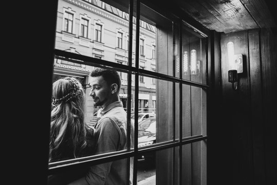 An interesting black and white photo on which a couple in love stands outdoors near a window of a cafe