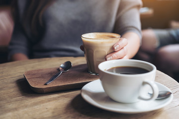 Closeup image of Americano coffee cup and woman's hand holding latte coffee cup on vintage wooden table in cafe
