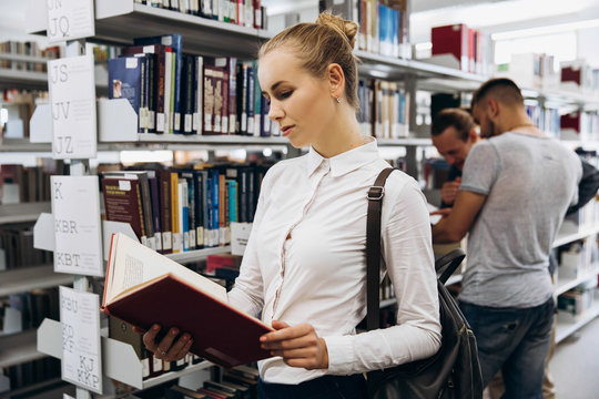 Pretty thoughtful girl looks like student standing with book in the library of a university