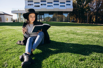 Stylish girl in black hat and coat sits with book and coffee on the lawn before the library