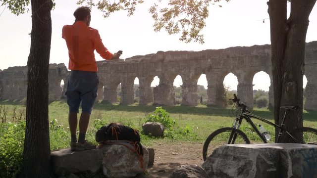 Cyclist with bike sitting on a rock under a tree taking selfies in front of ancient roman aqueduct. Young attractive athletic man with orange sportswear and backpack in parco degli acquedotti in Rome