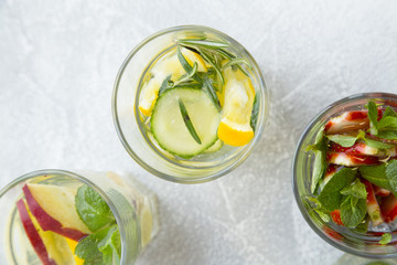 Three glasses of detox infused water with sliced strawberry, apples, lemons, cucumbers, mint and rosemary on light concrete background, top view