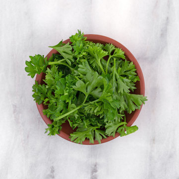 Top view of a red clay bowl filled with chopped curly parsley on a gray marble cutting board.