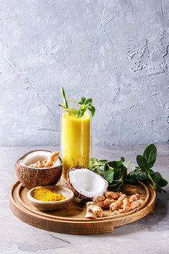 Cup of ayurvedic drink golden coconut milk turmeric iced latte with curcuma powder, crushed ice, mint and ingredients above on round wooden tray over grey kitchen table. Copy space