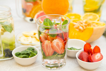 Glasses of detox water with sliced fruit, vegetables, berries and herbs and bowls with ingredients: strawberry, mint, spinach, grapefruit on a light stone background, selective focus, top view