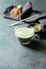 Cup of matcha latte with bowl of matcha tea and bamboo tools over grey green texture background....