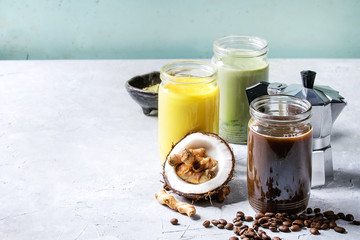Variety of iced colorful latte drinks. Iced coffee, turmeric and matcha latte cocktails in glass jars with ingredients above over grey green texture background. Copy space