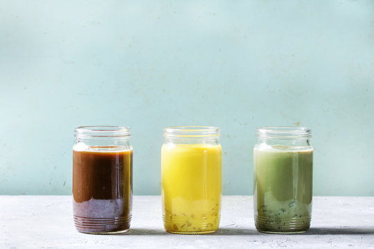 Variety of iced colorful latte drinks. Iced coffee, turmeric and matcha latte cocktails in glass jars over grey green texture background. Copy space