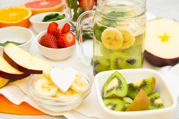 A glass jar of detox water with sliced banana, kiwi and mint and bowls with ingridients: banana, strawberry, kiwi, spinach and apple on a table with orange plastic cutting board