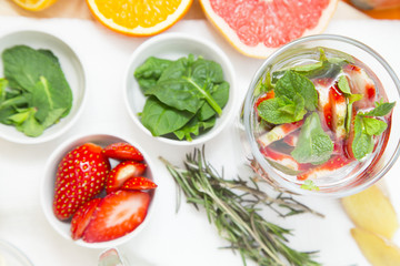 A glass of detox water with sliced strawberry and mint, cups with ingredients and a spring of rosemary, white background, top view