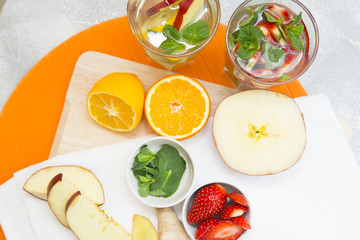 Detox infused water with slices of fruit and herbs.Bowls with strawberry and spinach, orange and lemon cut in halves, sliced apple and ginger on cutting board and white napkin, top view