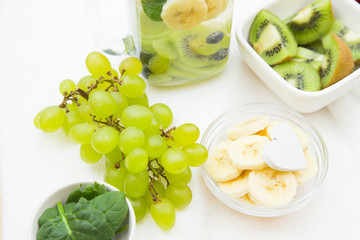 A jar of detox water with fresh fruit and ingredients on the table: green grapes, chopped kiwi, sliced banana and spinach, light stone and white napkin as a background, top view