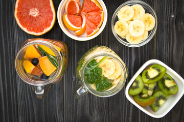 Glass jars and a glass of detox water with fresh fruit and herbs and ingredients on the table: grapefruit, chopped kiwi and slices banana in bowls, black wooden background, top view