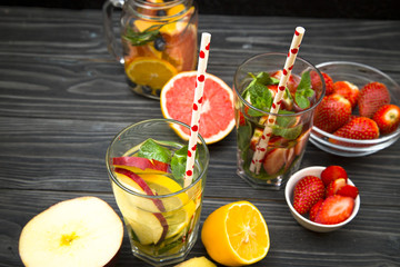 A jar and two glasses of detox water with straws with ingredients on black wooden table: fresh strawberry, ginger, apple, grapefruit and lemon cut in half