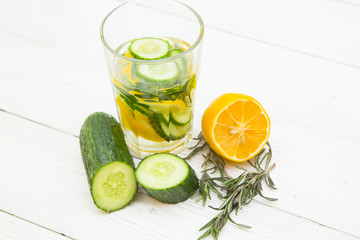 A glass of detox water with lemon, cucumber and a spring of rosemary and ingredients on the table, white wooden background