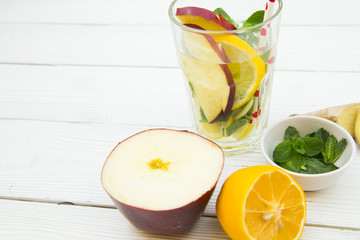 A glass of detox water with sleces of fresh apple, lemin ginges and spinach with ingredients on white wooden table