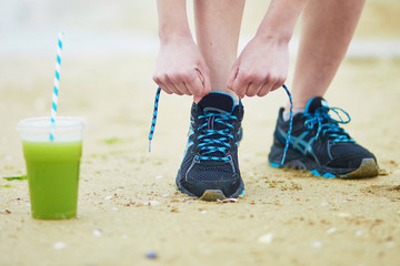 Jogger with green vegetable smoothie, tying sport running shoes laces