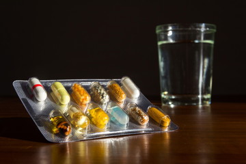 Detail of various colorful pills in transparent medicine capsule with glass of water on brown...