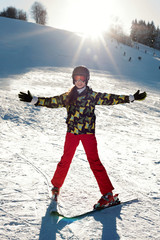 Fototapeta na wymiar A girl skier in red pants and a green jacket on skis stretching out her arms happy with the sun's rays