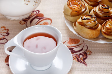 A cup of fruit apple tea with a saucer with baked rolls on a plate and a teapot on a napkin.