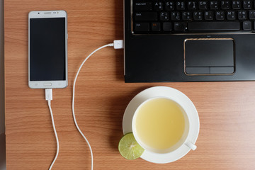 Top view, Plug in USB cord charger of the mobile phone with a laptop and freshly Lime juice in a white cup, on wooden floor