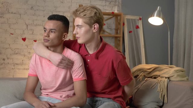 Pair of young multinational homosexuals sit on the couch, an American guy presents a gift to his partner. Homeliness, gay, young LGBT family concept. 60 fps