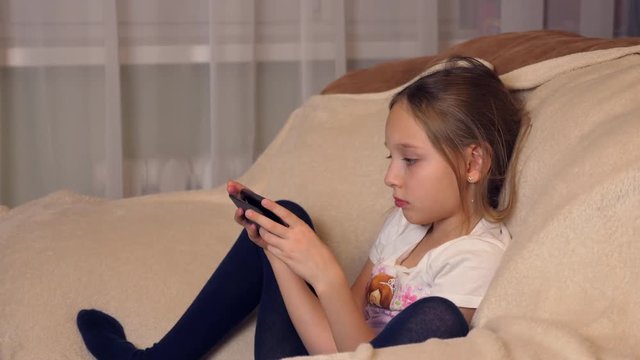 Little girl using modern smartphone sitting in soft chair at home.