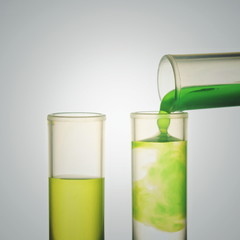 Scientist hand pouring a green chemical solution from a laboratory glass test tube into a scientific cylinder with yellow liquid for an experiment in a science research lab