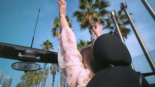 Back shot of unrecognizable happy girl in convertible car rises her hands and swinging it side to side isolated on blue sky with palm trees passing by. Freedom and happiness concept.