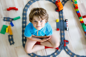 Fototapeta na wymiar little blond kid boy playing with colorful plastic blocks and creating train station