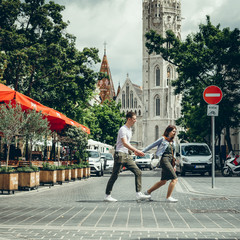 The full-length view of the loving couple of tourists holding hands while crossing the road at the background of St. Matthew's Cathedral in Budapest, Hungary.
