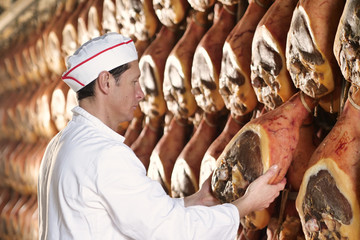 In a ham factory, a man in charge of quality control walks between the hams and controls, the perfume and the certified Italian quality. Concept of: tradition, Italy, food, ham - obrazy, fototapety, plakaty