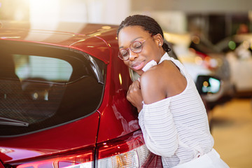 Smiling african woman hugging a red car at new car showroom