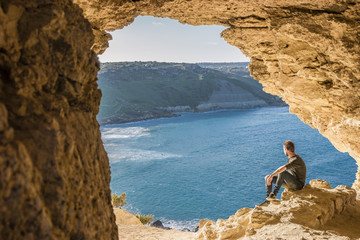 young man standing in a cave looking out over the ocean by Tal Mixta Cave Malta Island Europe ,Gozo Malta - obrazy, fototapety, plakaty